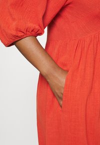 Robe en lin rouge avec des manches bouffantes et un corsage ajusté. Elle présente une poche latérale, un tissu texturé et des détails de jupe froncée.