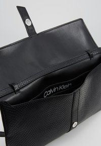 Černá kabelka značky Calvin Klein otevřená, ukazuje texturovaný vnější povrch, hladký vnitřek a magnetický uzávěr.