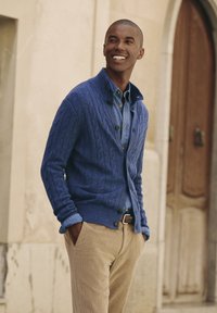 Blauw kabelgebreid vest over een denim overhemd, gecombineerd met lichtbeige corduroypantalon. Heeft knopen en een sjaalkraagontwerp.