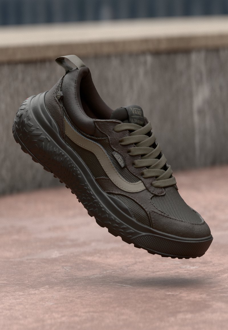 Dark brown sneaker with textured fabric, leather accents, green laces, and a rugged rubber sole. Features branding on the heel and tongue.