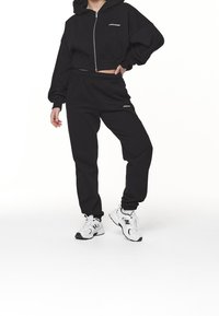 Sudadera corta negra con bolsillo frontal, cierre de cremallera y logo. Pantalones de chándal negros con cintura elástica y logo. Zapatillas blancas.