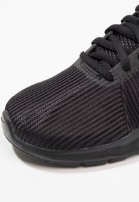 Black athletic sneaker with a textured upper featuring vertical stripes, round toe, and black laces. Soles have a smooth finish.