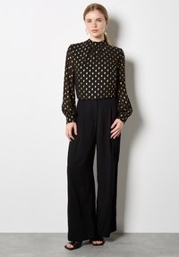Schwarze Bluse mit langen Ärmeln und goldfarbenem Blattmuster, hochgeschlossen und mit gerafften Details, kombiniert mit schwarzen weitgeschnittenen Hosen und offenen Absatzschuhen.