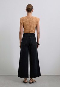 Beige strukturierte Bluse mit kurzen Ärmeln, kombiniert mit hochtaillierten, weiten, dunklen Jeans und schwarzen Riemchensandalen, von hinten betrachtet.