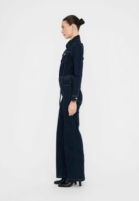 Giubbotto in denim blu scuro con maniche lunghe, parte superiore aderente, fondo a gamba larga e dettagli in bottone argento. Indossato con stivaletti neri con tacco.
