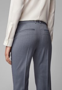 Grijs geruit pantalon met een gladde textuur, met twee achterzakken, gecombineerd met een lichtgekleurde blouse, van achteruit gefotografeerd.