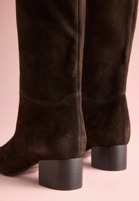 Zwarte suède kniehoge laarzen in donkerbruin met een slank ontwerp, voorzien van een ronde neus en een chunky zwarte hak. Gladde textuur en minimale stiksels.