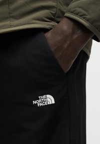 Δεξί χέρι σε τσέπη μαύρου παντελονιού με λευκό λογότυπο "The North Face", φοράει ελιά πράσινο μπουφάν με ανεστραμμένο μανίκι.