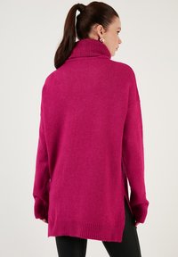 Fuksia neulotun villatakin, jossa on korkea kaulus, pitkät hihat ja sivupitsit. Pehmeä pinta ja ribbivarat kauluksessa ja helmassa.