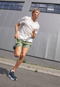 Pantaloni sportivi color verde oliva, in tessuto leggero. T-shirt bianca con un piccolo logo. Scarpe da corsa in blu scuro con suola bianca. Sfondo urbano.