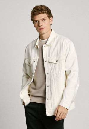 Veste en denim blanc avec des boutons en métal, deux poches poitrine et des détails de couture subtils, superposée à un pull beige.