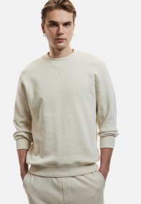 Sudadera beige clara de tejido suave, con cuello redondo, puños y dobladillo acanalados, y un sutil patrón texturizado en los hombros.