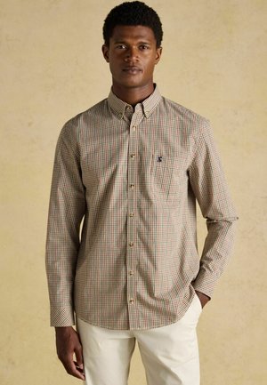 Hemd mit Button-Down-Kragen in einem karierten Muster aus Beige, Grün und Rot. Lange Ärmel, Kragen und ein kleines Brustlogo. Getragen mit hellen Hosen.