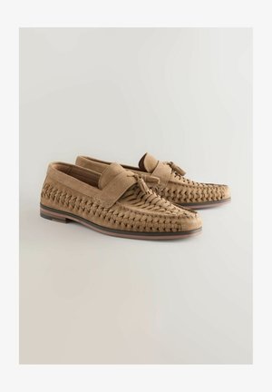 Beige Wildleder Slipper mit einem gewobenen Design, flacher Sohle und einem Fransenakzent an der Vorderseite. Der Schuh hat ein glattes Innenfutter und eine braune Umrandung.