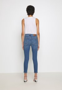 Úzké modré denimové džíny s středním pasem, pětikorunovým designem a hnědou koženou nášivkou na zadní straně. Hladká textura, přiléhavý střih.