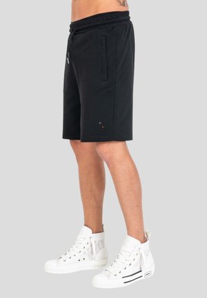 Uomo che indossa pantaloncini neri fino al ginocchio con tasche laterali e sneakers alte bianche su uno sfondo semplice.