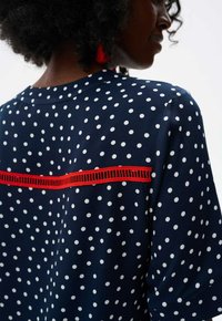 Marineblauwe blouse met witte stippen, voorzien van een rode horizontale accentstreep en een gestructureerd detail op de achterkant. Korte mouwen, soepele stof.