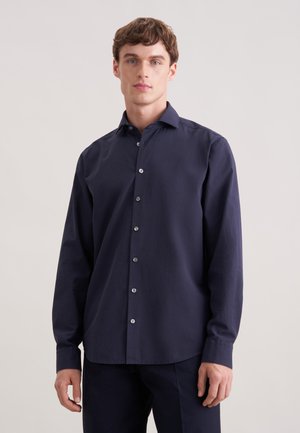 Ung mand, der står iført en mørk navy blå skjorte med lange ærmer og matchende bukser mod en ensfarvet lys baggrund.