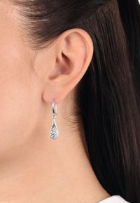 Silver teardrop-örhängen med en texturerad design, som har en hengslet ring och glittrande accenter, bärs på en models öra.