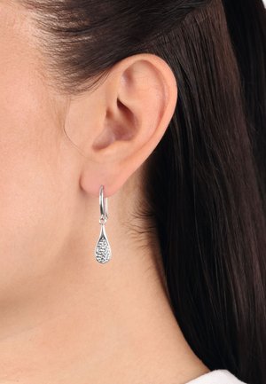 Orecchini a goccia in argento con un design testurizzato, caratterizzati da un anello a cerniera e accenti scintillanti, indossati all'orecchio di un modello.