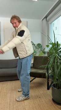 Fyldig beige jakke med en sort lap på venstre ærme, båret over en sort skjorte, kombineret med lyseblå jeans og grå sneakers.