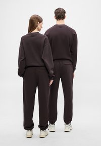 Tmavohnedé mikiny a tepláky s rebrovanými manžetami a voľným strihom, s jemným logom na zadnej strane, vyrobené z mäkkej tkaniny.