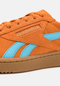 Reebok tenisky v oranžové semišové barvě s modrými syntetickými pruhy, s texturovanou gumovou podrážkou a zlatým logem na boku.