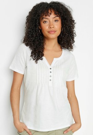 Mujer con cabello rizado, usa pendientes de aro y una camisa blanca de manga corta con botones y plisados verticales, de pie con las manos en los bolsillos.