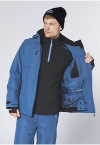 Blaue isolierte Jacke mit durchgehendem Reißverschluss und Kapuze, verfügt über eine Brusttasche, schwarzes Innenfutter und hellblaue Reißverschlüsse. Kombiniert mit einem schwarzen Hemd.