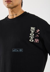 T-shirt en coton noir avec impression multicolore en texte japonais et anglais sur le devant et une étiquette bleue près de l'ourlet.