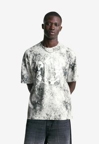 Junger Mann in einem T-Shirt mit weiß-schwarzem abstraktem Muster und erhabenem weißen Runddesign, kombiniert mit dunkel gewaschenen Jeans, stehend mit Händen hinter dem Körper.