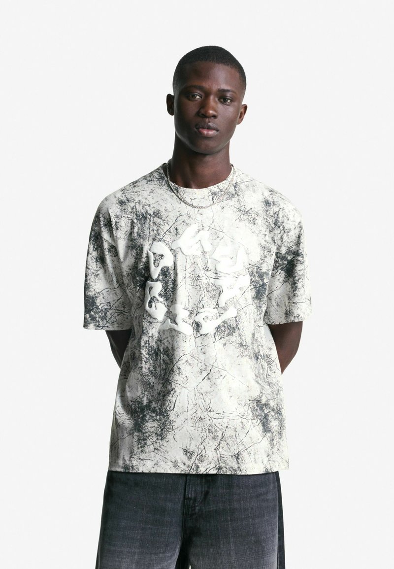 Junger Mann in einem T-Shirt mit weiß-schwarzem abstraktem Muster und erhabenem weißen Runddesign, kombiniert mit dunkel gewaschenen Jeans, stehend mit Händen hinter dem Körper.