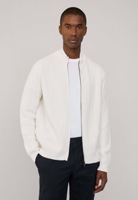 Cardigan bianco a coste con colletto alto e zip completa, dotato di maniche lunghe e vestibilità morbida, indossato sopra una camicia bianca.