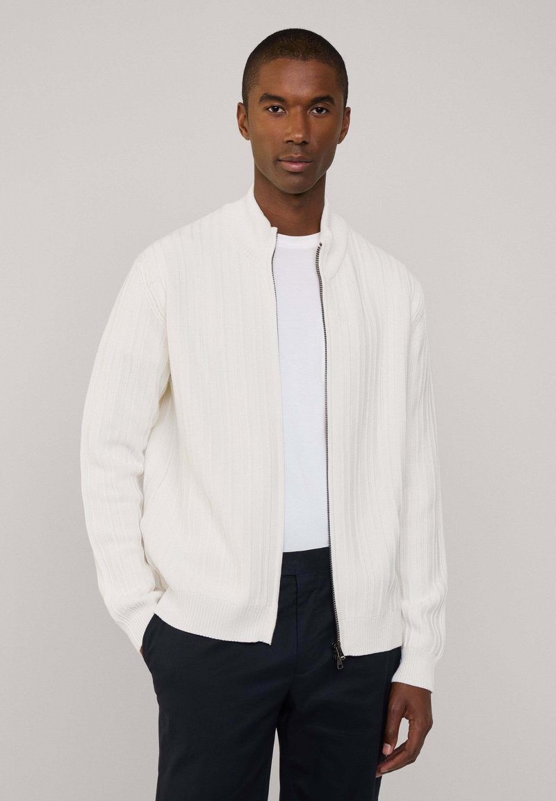 Cardigan bianco a coste con colletto alto e zip completa, dotato di maniche lunghe e vestibilità morbida, indossato sopra una camicia bianca.