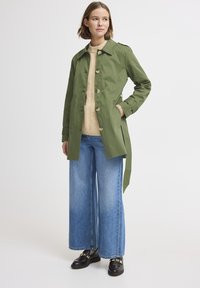 Cappotto verde oliva con colletto, chiusura a bottoni e tasche laterali, indossato sopra un maglione beige e jeans a gamba larga blu. Scarpe nere con dettagli.