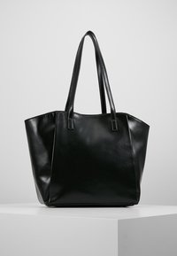 Black leather tote bag with two shoulder straps standing upright on a white pedestal against a plain gray background.