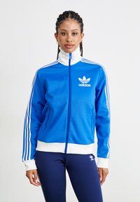 Mėlynas užtrauktuku užsegamas džemperis su baltomis juostelėmis ir Adidas logo. Pagamintas iš lygaus audinio, turi aukštą apykaklę ir šonines kišenes. Suderintas su tamsiai mėlynomis leginsais.