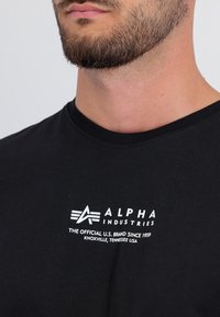 Mustmust shirt, millemust ja valge trükitud logo ja tekstiga rinnal. Krae on standardne ja kangas tundub sujuv.