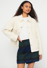 Kreemikas fliisjope kraega, snap-nuppude ja esitaskuga, sobitatud tume rohe-rohelise ja sinise ruudulise miniseelikuga.