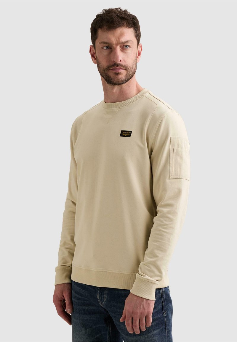 Homme portant un sweat-shirt beige à manches longues avec un petit patch logo sur la poitrine et une poche sur la manche gauche, associé à un jean bleu.