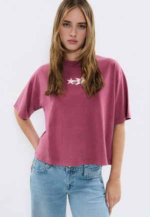 Mujer joven con cabello rubio largo que lleva una camiseta amplia color malva con un logo de estrella blanca y jeans azul claro, de pie frente a un fondo liso.