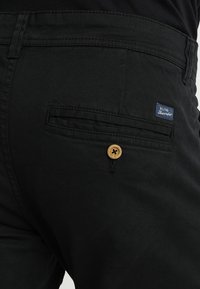 Primer plano del bolsillo trasero de unos pantalones negros con cierre de botón y una pequeña etiqueta azul "Blend Headrmaker" encima del bolsillo.