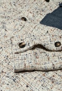 Približni prikaz bež tkanine teksture s šarenim mrljama, prikazujući presavijeni rub i tamne okrugle gumbe.