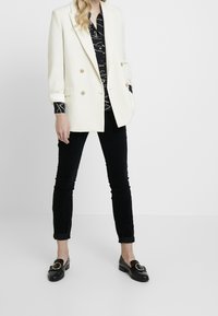 Bílý dvouřadý blazer se zlatými knoflíky, zkombinovaný s černou vzorovanou košilí a černými slim-fit kalhotami. Černé mokasíny s kulatým detailem.