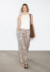 Witte mouwloze top, beige broek met bruine gespikkelde patronen en een bruine schoudertas. Model staat op een grijze vloer tegen een witte muur.