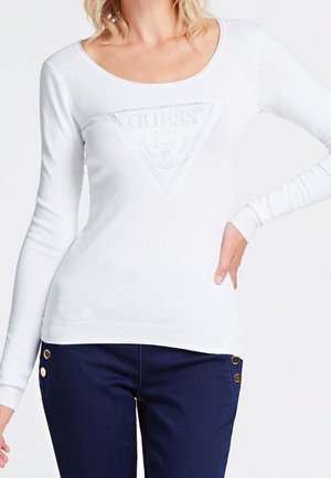 Kvinde iført en hvid langærmet skjorte med et Guess-logo og mørkeblå bukser med guldknapper, hånd på hoften.