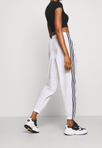 Pantalon de jogging blanc avec bandes latérales noires, taille élastique et chevilles resserrées, accompagné de baskets noires et blanches ainsi qu'un crop top noir.