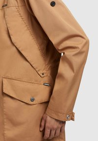 Sandfarbene wasserdichte Jacke mit einer Taschenklappe, Druckknöpfen und einer eleganten, glatten Textur. Verfügt über eine Schulternaht und ein markenspezifisches Etikett.