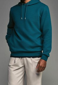 Turkusowy, teksturowany hoodie z kapturem ze sznurkiem, ściągaczami przy mankietach i u dołu, w połączeniu z jasnobeżowymi spodniami. Cechuje się luźnym krojem i przednią kieszenią.