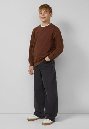 Sweat-shirt marron à manches longues avec poignets côtelés, pantalon noir à jambes larges, et chaussures blanches avec accents beige. Le mannequin sourit et est debout.